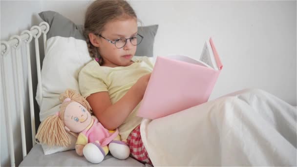 Kislány az üvegek, miközben az ágyban fekve egy könyvet olvas. Mellette ül a baba. — Stock videók