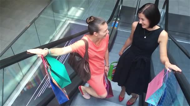 Dos chicas moviéndose arriba en escaleras mecánicas en el centro comercial, amigos están de compras en una tienda — Vídeos de Stock