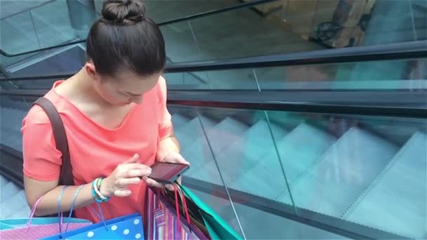 Jovem mulher usando smartphone na escada rolante no shopping center e usando telefone inteligente. Estilo de vida urbano, mantendo contato a cada minuto — Vídeo de Stock