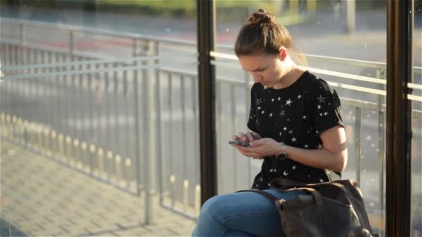 Jovem mulher caucasiana encantadora com olhar moderno usar telefone celular enquanto espera transporte na parada de ônibus, mulher segurando telefone celular enquanto sentado em uma estação contra o pôr do sol fundo — Vídeo de Stock