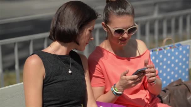 Chicas hermosas jóvenes usando teléfono inteligente sentado en la parada de autobús de la ciudad, Dos amigos hembra riendo en el banco — Vídeo de stock