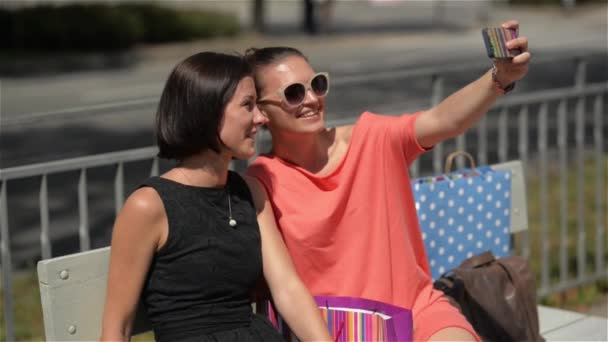 Dos chicas están haciendo selfie con el teléfono después de ir de compras, hermosa mujer joven sentada en un banco de la ciudad y riendo — Vídeos de Stock