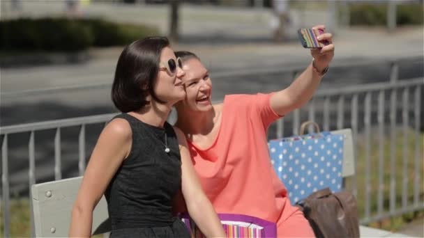 İki kız şehirde bir bankta oturmuş ve gülerek alışveriş, güzel genç bir kadın sonra Selfie telefon ile yapıyoruz — Stok video