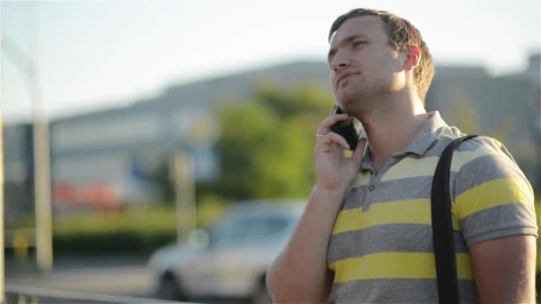 Молодой человек держит мобильный телефон, использует смартфон, делает звонок, разговаривает по телефону, стоит на солнечной улице с транспортным движением на заднем плане — стоковое видео