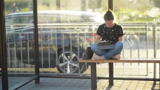 Junge charmante Kaukasierin mit trendigem Look benutzt Tablet während des Wartens Transport an Bushaltestelle, Frau sitzt auf einem Bahnhof vor Sonnenuntergang Hintergrund — Stockvideo