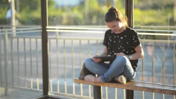 Νεαρή γυναίκα διαβάζοντας ένα Tablet ή e-Book σε έναν σιδηροδρομικό σταθμό, ενώ είναι σε αναμονή για τις δημόσιες μεταφορές και κοιτάζοντας το της ρολόι — Αρχείο Βίντεο