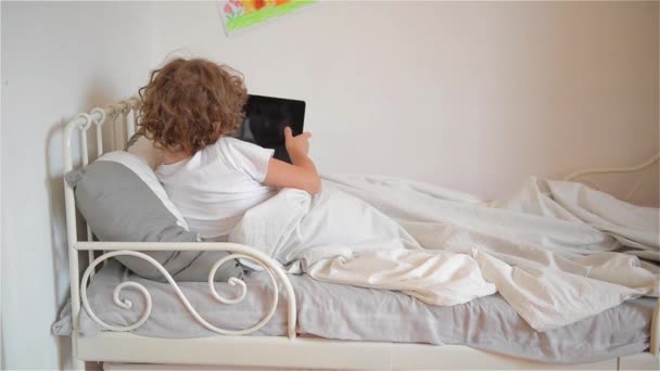 Χαριτωμένο μικρό αγόρι που είναι ξεκούραστη στο κρεβάτι του, παίζοντας παιχνίδια στον υπολογιστή ψηφιακή δισκίο — Αρχείο Βίντεο