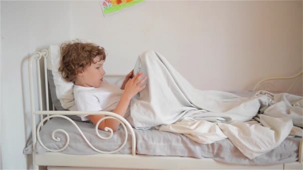 Милый мальчик расслабляется на кровати, играя в компьютерные игры на планшете — стоковое видео