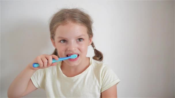 Linda niña con coletas que se cepillan diligentemente los dientes. En la mano de la chica tiene cepillo de dientes azules. alegre chica mirando en la cámara . — Vídeo de stock