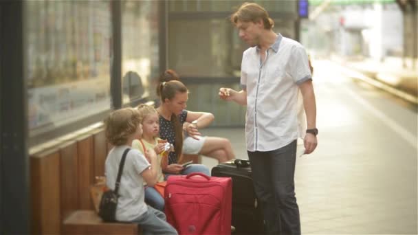 Lycklig familj väntar hans tåg vid järnvägsstationen och äta på en bänk, ser mor på klockan — Stockvideo
