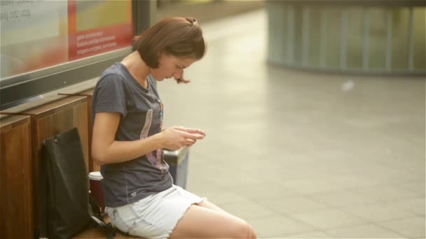 Ung kvinna sitter i väntar på bänken i järnvägsstationen, hålla smartphone, tittar på skärmen, använda mobiltelefon app, att skriva eller läsa textmeddelande — Stockvideo