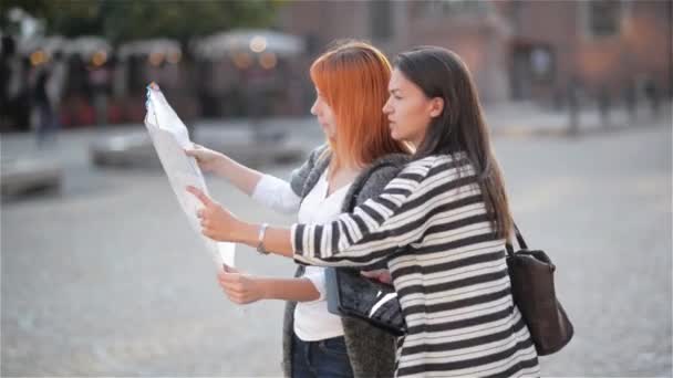 Två unga vackra flickor tittar åt sidan i den urbana zonen medan du utforskar staden. Kaukasiska kvinnor vandrar med papper kartan för att hitta intressanta platser när du reser. — Stockvideo