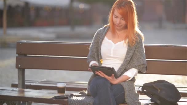 Молодая женщина с рыжими волосами сидит на деревянной скамейке в городе и с помощью планшетного ПК, студентка читает электронную книгу, старый город фон — стоковое видео