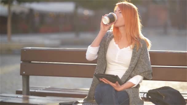 Mädchen mit roten Haaren sitzen auf einer Bank an der Straße und trinken Kaffee, junge Frau mit Tablette an einem sonnigen Tag, Hintergrund der Stadt — Stockvideo