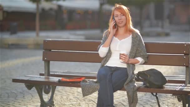 Mulher bonita com cabelo vermelho falando no telefone celular sentado em um banco na rua com edifícios no fundo, menina bebendo café e rindo — Vídeo de Stock