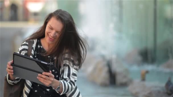 Joyeux jeune femme avec tablette informatique ayant chat vidéo tout en étant assis sur le banc à la rue, fond de fontaine — Video