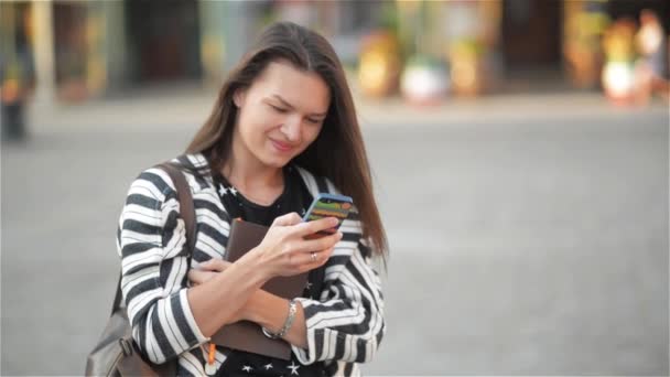 Красивая молодая женщина с помощью смартфона на городской улице, студентка держит телефон и ноутбук, старый город — стоковое видео