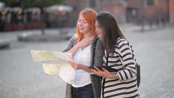 Due giovani ragazze per le strade della città vecchia. Le fidanzate cercano di trovare la loro strada in una città sconosciuta. Ragazza dai capelli rossi che guarda le mappe. Il suo compagno di viaggio dai capelli scuri guarda sullo schermo del tablet — Video Stock