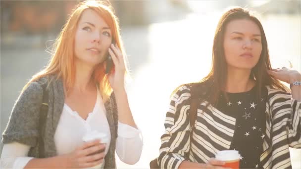 Twee schattige meisjes lopen de straat in de stad. Meisjes houden papieren bekers met koffie. Roodharige meisje spreekt op een mobiele telefoon. Haar vriend ziet er op zijn horloge en drinken koffie — Stockvideo