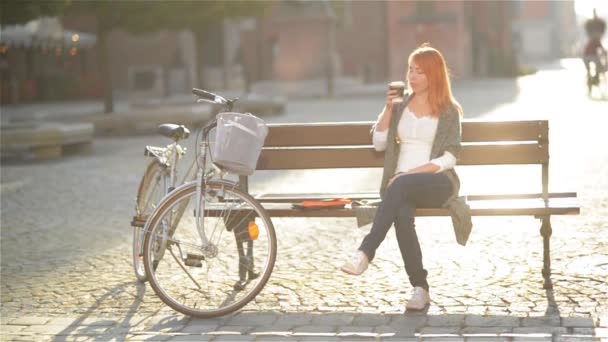 Cute redheaded girl is resting, sitting on a bench in the city. She postponed aside her Tablet and drink coffee — Stock Video