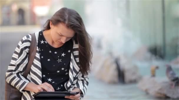 Hermosa chica leyendo y hojeando un libro electrónico o una tableta sentada en un banco en la calle, fondo de fuente — Vídeo de stock