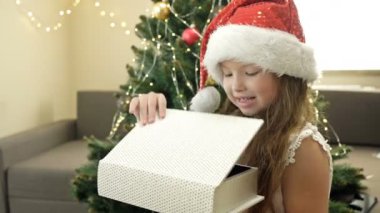 Noel ağacının arka planında Noel Baba şapkalı mutlu küçük kız. Çocuk Noel hediyesiyle mutlu..
