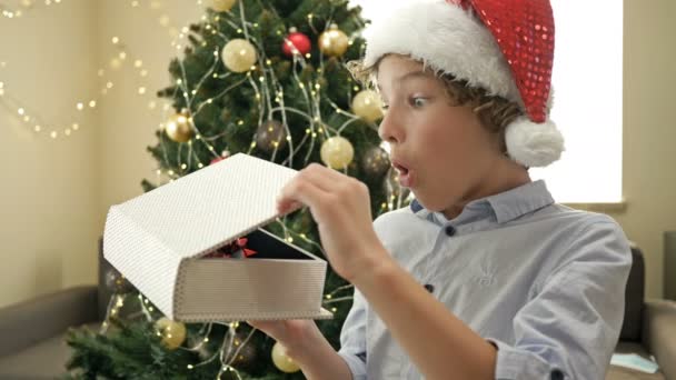 Lykkelig teenage dreng i Santa hat på baggrund af juletræet. Youngster er tilfreds med julegaven. – Stock-video