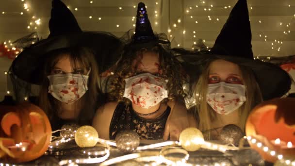 Три девочки-подростки в черных костюмах, ведьмы в медицинских масках, сидят за столом. Хэллоуин атрибуты на столе. Хэллоуин во время пандемии коронавируса covid19. — стоковое видео