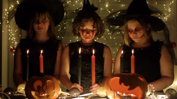Tres chicas adolescentes disfrazadas de brujas con una mirada aterradora en sus rostros apagando velas. Fondo oscuro. Atributos de Halloween sobre la mesa. — Vídeo de stock
