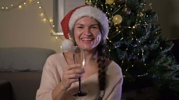 Schöne, fröhliche Frau mit Weihnachtsmütze und einem Glas Champagner. Vor dem Hintergrund eines Weihnachtsbaums. Frohes Neues Jahr und frohe Weihnachten. — Stockvideo