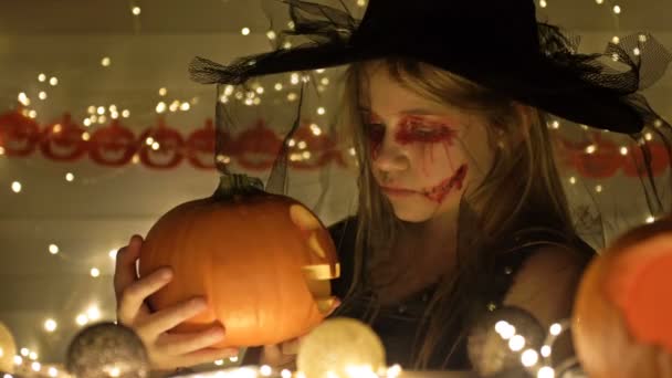Porträtt av en tonårstjej klädd som en häxa med skrämmande smink i ansiktet. Flickan håller en Jack-o-lantern i sina händer. Mörkt guld bakgrund. Halloween. — Stockvideo