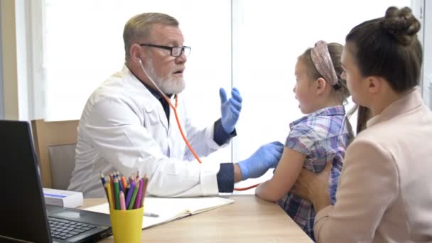 Petite fille avec sa mère à un rendez-vous chez le pédiatre. Un pédiatre masculin âgé examine un enfant dans son bureau, écoute les poumons ou le cœur des enfants avec un phonendoscope. — Video