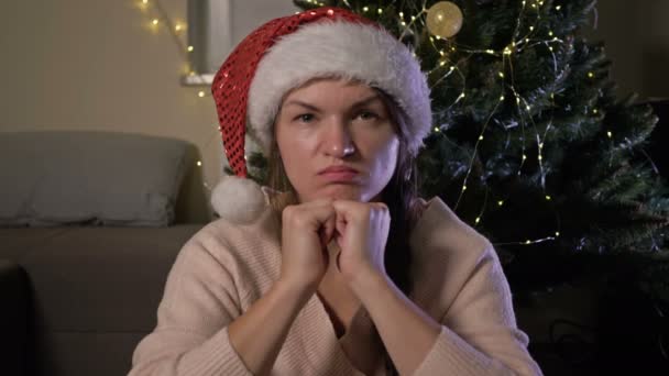 美しいです中世の女性で赤いパーティーサンタ帽子座っていますザクリスマスツリーの近くに悲しい表情で顔. — ストック動画