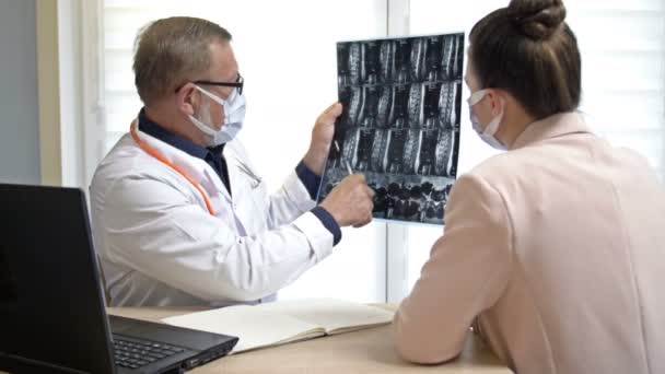 Senior male traumatologist explaining x-rays to the female patient. Doctor and patient in medical masks. Covid-19. — Stock Video