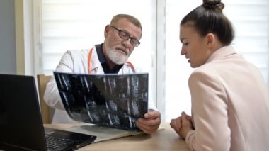 Deneyimli bir erkek doktor röntgen görüntüsü gösterir ve bir tıp merkezinde muayene sırasında genç bir kadın hastaya teşhisi açıklar..
