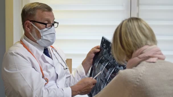Traumatólogo masculino mayor explicando radiografías a la paciente femenina. Médico y paciente con máscaras médicas. Covid-19. — Vídeo de stock