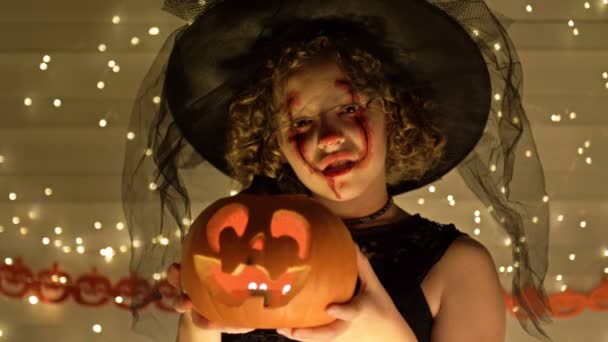 Portrét dospívající dívky oblečené jako čarodějnice s děsivým make-upem na tváři. Dívka drží v rukou Jack-o-lucernu. Tmavě zlaté pozadí. Halloween. — Stock video
