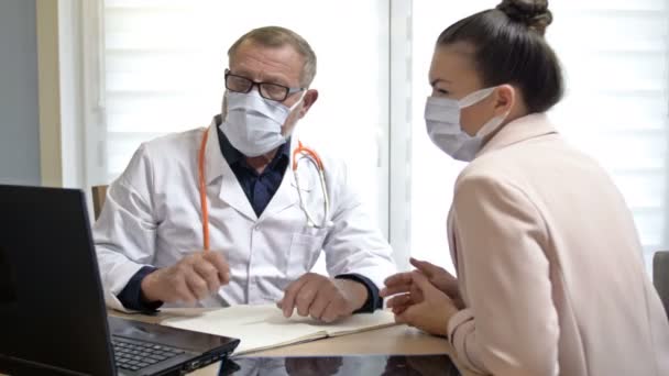 Deneyimli bir erkek doktor kadın bir hastaya danışır. Doktor ve hasta tıbbi maske takıyor. COVID-19 salgını sırasında harekete geçilmeli. — Stok video
