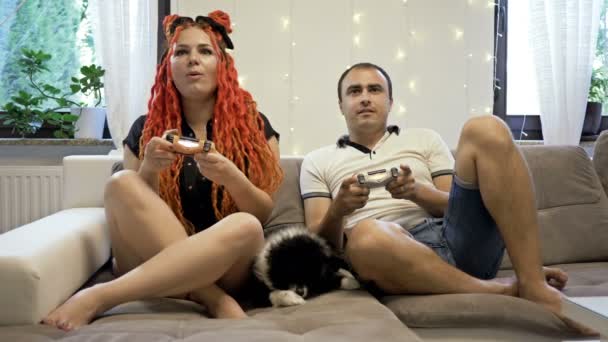Pareja casada joven está jugando juegos de ordenador en casa. Un perro pequeño está acostado en el sofá cercano. Pasar tiempo juntos. — Vídeos de Stock