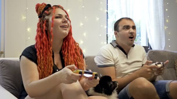 Młode małżeństwo gra w gry komputerowe w domu. Mały piesek leży na pobliskiej kanapie. Spędzać razem czas. — Wideo stockowe