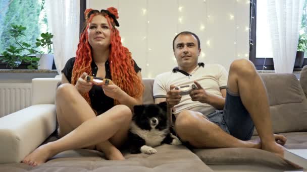 Jong getrouwd stel speelt computerspelletjes thuis. In de buurt ligt een kleine hond op de bank. Samen tijd doorbrengen.. — Stockvideo
