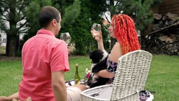 Un par de amantes tuvieron un romántico picnic en el patio de su casa. Ambos están bebiendo vino.La mujer sostiene un pequeño perro lindo en sus manos. — Vídeo de stock