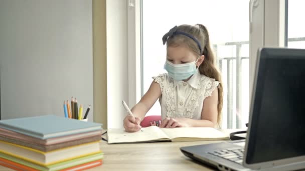A kis iskoláslány orvosi maszkban épp a házi feladatát csinálja. Tanulás a COVID-19 járvány során. — Stock videók
