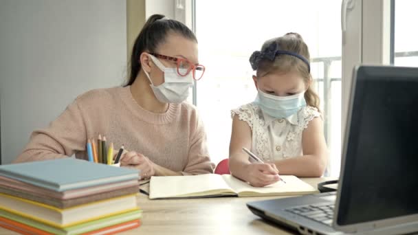 Mom helps her little daughter with lessons. Both have medical masks. Distance learning ar during the COVID-19 pandemic. — Stock Video