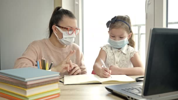 Moeder of leraar of mentor helpt een basisscholier met haar lessen. Beide hebben medische maskers. Training tijdens de COVID-19 pandemie. — Stockvideo