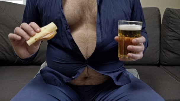 L'uomo è seduto sul divano, mangiando un panino e bevendo birra da un grande bicchiere. La sua camicia non è abbottonata a causa della pancia enorme. — Video Stock