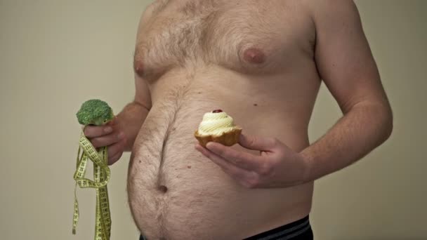 Şişman adam bir elinde brokoli, diğer elinde pasta tutuyor. Sağlıklı ve sağlıksız yiyecekler arasında zor bir seçim.. — Stok video