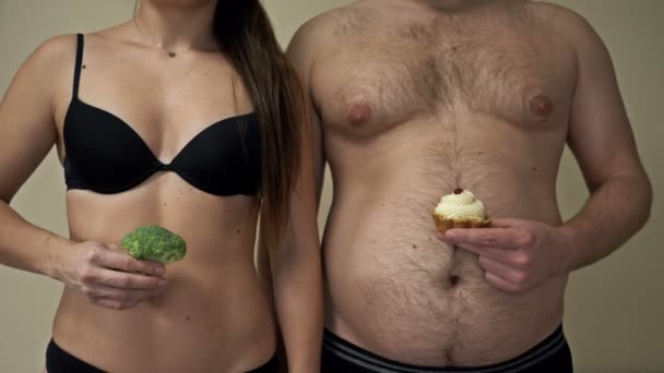 Mujer delgada sostiene brócoli en su mano, y un hombre gordo sostiene un pastel. Las consecuencias de una dieta saludable y poco saludable. — Vídeo de stock