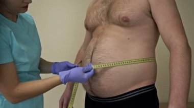 Doktor, tombul bir erkek hastanın karın çevresini ölçer. Aşırı kilolu problemler.