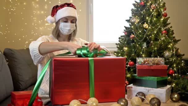 Hermosa mujer con un sombrero de Santa Claus y una máscara protectora en su cara está empacando regalos. En el fondo del árbol de Navidad. Navidad a pesar de la pandemia de COVID-19. — Vídeos de Stock